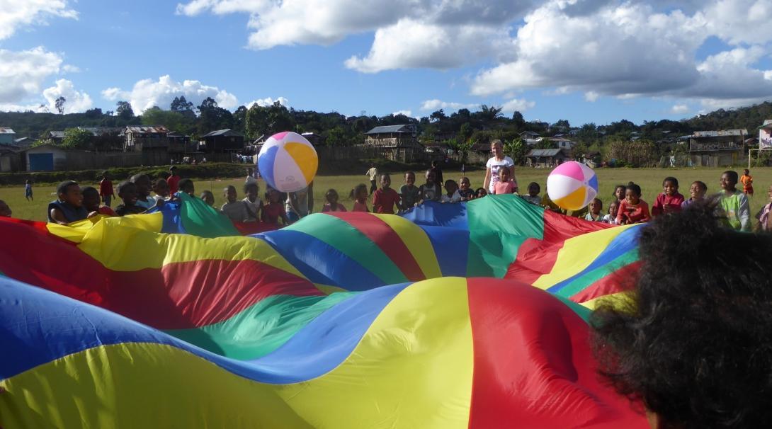 Kinderopvang vrijwilligers spelen een spel met kinderen met een grote parachute op de school in Madagaskar. Projects Abroad vrijwilligers werken met kinderen in Madagaskar en spelen buitenactivietiten op een lokale school met een grote parachute die helpt met het ontwikkelen van motorische vaardigheden.